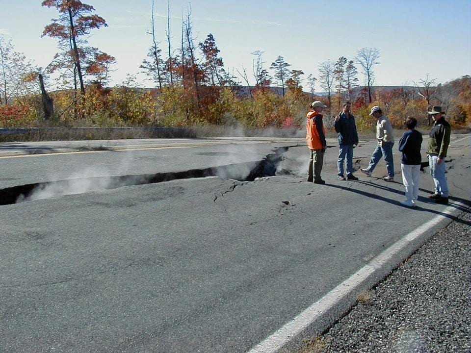 A two-lane asphalt highway split by a long crack, with white vapour rising from the fissure into cold sunlight. Five people in coats stand on the intact side of the road looking at it. Bare and orange-leaved trees line the background under a clear blue sky.